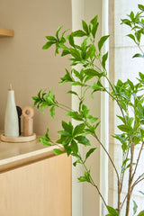 Een kunstplant Pieris Japonica van 150 cm staat bij een raam naast een licht houten kastje, waarop keramische flesjes en een pepermolen staan, wat een minimalistische toets geeft aan de lichte, luchtige kamer.