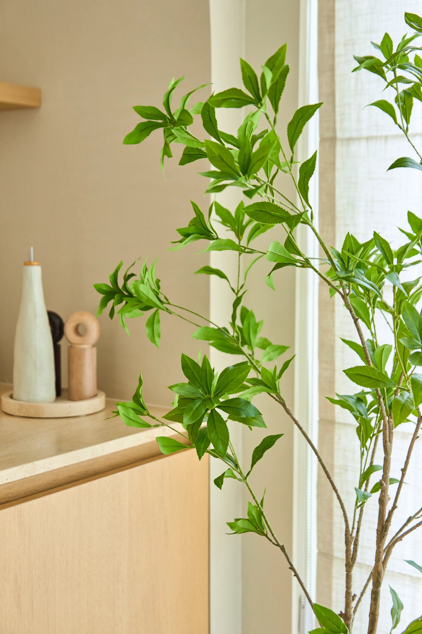 Een kunstplant Pieris Japonica van 150 cm staat bij een raam naast een licht houten kastje, waarop keramische flesjes en een pepermolen staan, wat een minimalistische toets geeft aan de lichte, luchtige kamer.