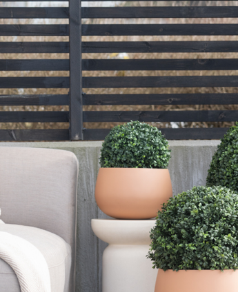 Drie Kunstplant Buxus Romeinse Stijl UV D30cm ronde topiaries staan op een patio bij een beige buitenstoel, tegen een modern zwart latten hek en kale bomen.