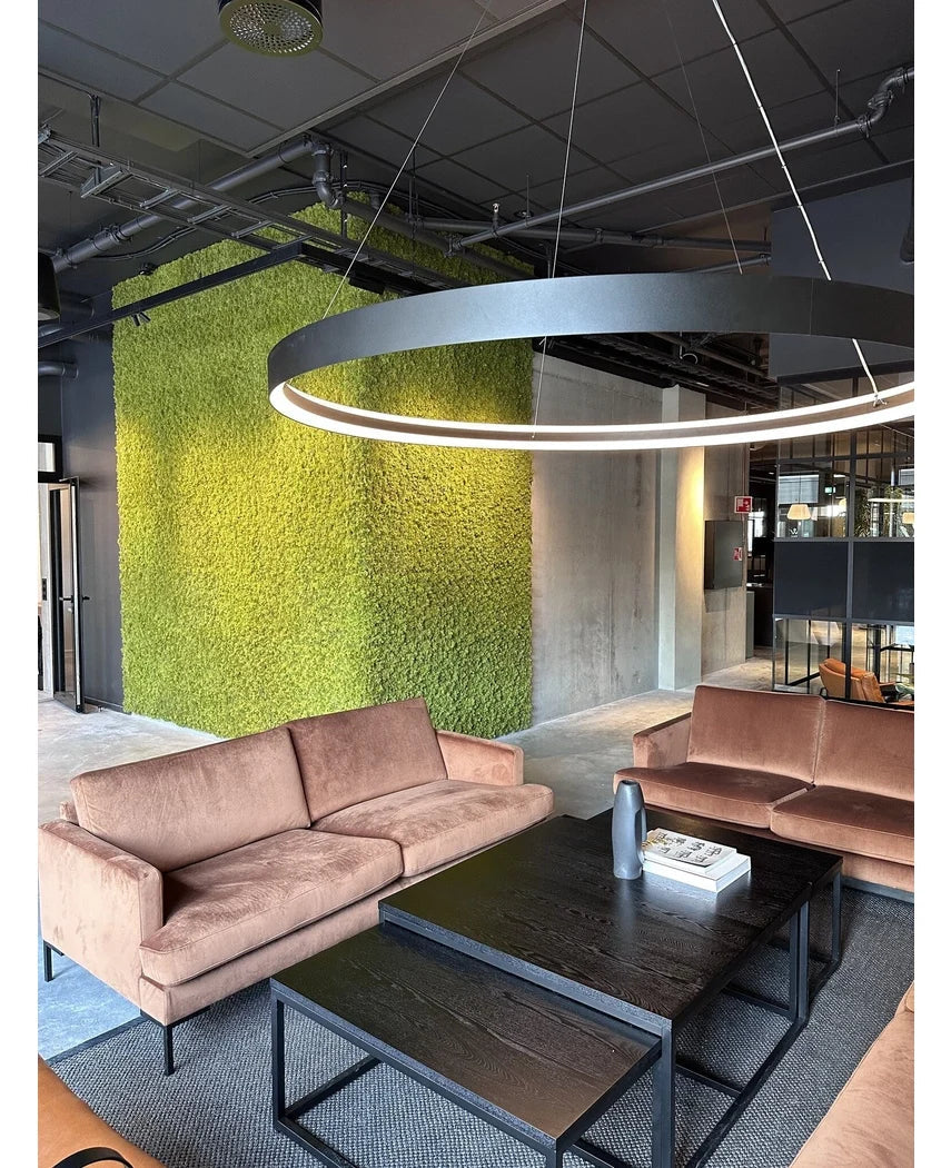 Et moderne opholdsområde med beige sofaer, sorte sofaborde, en stor rund loftslampe og en grøn væg i baggrunden, skabt med Kunsthaag Nordic Moss 50x50 cm UV, som udstråler et luksuriøst interiør og et moderne industrielt design.