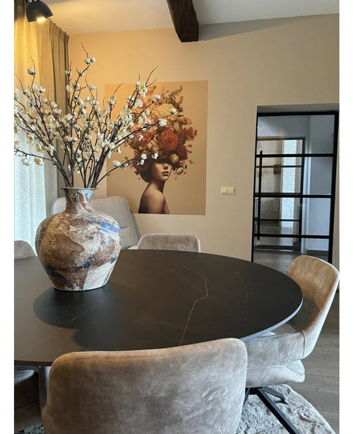 En moderne spiseplads med et rundt sort bord, beige stole og en stor keramikvase med kunstig japansk kirsebærblomst 94 cm hvid. Et vægmaleri af en kvinde med blomster i håret dekorerer baggrunden.