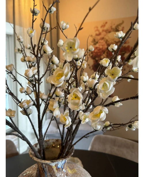 Et nærbillede af den kunstige japanske kirsebærblomst 94 cm med yndefulde hvide kunstige kirsebærblomster med gule centre på brune grene i en glasvase, på et mørkt bord med et elegant, blødt oplyst rum i baggrunden.