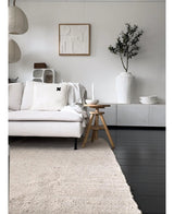 En minimalistisk stue med lys sofa, neutralt tæppe, hvid skænk, kunstigt oliventræ 120 cm som dekorativt element, abstrakt vægkunst og træskamler - alt sammen i en blød middelhavsatmosfære med neutrale toner.