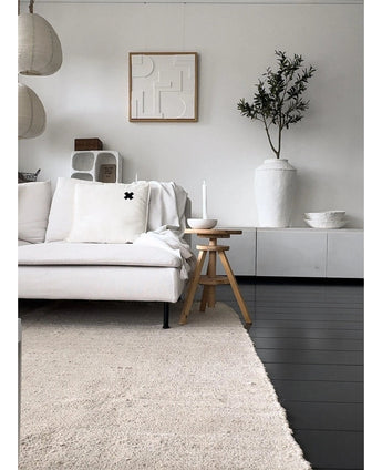 En minimalistisk stue med lys sofa, neutralt tæppe, hvid skænk, kunstigt oliventræ 120 cm som dekorativt element, abstrakt vægkunst og træskamler - alt sammen i en blød middelhavsatmosfære med neutrale toner.