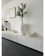 Et minimalistisk værelse med hvid skænk, det kunstige oliventræ 120 cm som dekorativt element, dekorative hvide skåle, et stearinlys og abstrakt vægkunst. Mørke trægulve og moderne accenter skaber en fredfyldt middelhavsatmosfære.