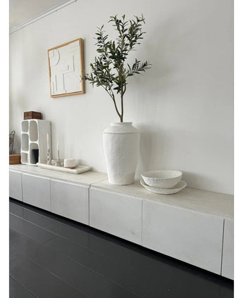 Et minimalistisk værelse med hvid skænk, det kunstige oliventræ 120 cm som dekorativt element, dekorative hvide skåle, et stearinlys og abstrakt vægkunst. Mørke trægulve og moderne accenter skaber en fredfyldt middelhavsatmosfære.