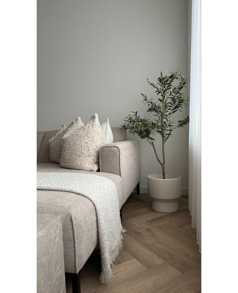 En moderne stue med en beige sofa, teksturerede hynder, et hvidt tæppe med frynser og et kunstigt oliventræ 120 cm som dekorativt element ved siden af ​​et vindue med hvide gardiner på et lyst gulv med sildebensmønster.