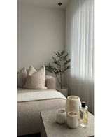 En minimalistisk stue har en beige sofa, teksturerede hynder, et kunstigt oliventræ 120 cm som indretning, gennemsigtige hvide gardiner og et marmorbord med tre cremefarvede vaser og parfume. Naturligt lys oplyser det rolige rum.