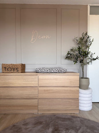 En minimalistisk planteskole indeholder en kommode i træ med pusleunderlag med leopardprint, en vævet kurv mærket "Legetøj" som dekoration, det kunstige oliventræ 120 cm for grønt og "Dean" i kursiv på den beige væg ovenfor.