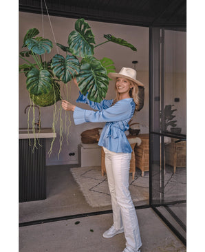 Een lachende vrouw met hoed, blauwe blouse en witte broek staat binnen en houdt een Kunst hangplant Monstera Deluxe 60 cm met grote groene bladeren vast, omringd door modern decor voor een tropische sfeer met decoratieve planten.