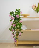 En træhylde med kunstig hængeplante Petunia 70 cm lilla, et hvidt ur, et træstativ, en bog og en vase med lyserøde blomster mod en hvid væg.