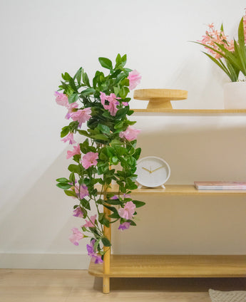 En træhylde med kunstig hængeplante Petunia 70 cm lilla, et hvidt ur, et træstativ, en bog og en vase med lyserøde blomster mod en hvid væg.