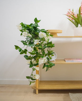 De Kunst hangplant Petunia 70 cm wit met witte bloemen valt van een houten schap in een lichte, moderne kamer met lichte houten vloeren. Op de bovenste plank staat een vaas met lange roze bloemen.