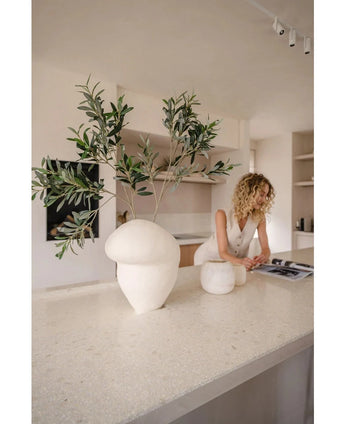 Een vrouw met krullend blond haar in een witte mouwloze outfit leunt over een tijdschrift op een licht aanrecht, op de voorgrond versierd met UV bestendige Kunst olijfblad 90 cm in moderne witte vazen.