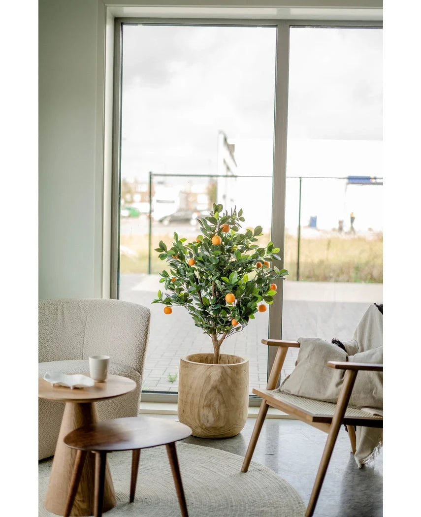 Een Kunst sinaasappelboom 120 cm staat bij een groot raam in een moderne, lichte woonkamer met een beige stoel, houten bijzettafel en een koffiekopje op een schoteltje.