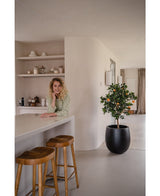 Een vrouw met krullend blond haar glimlacht, leunend op een aanrecht. Naast haar geven twee houten krukjes en een Kunst sinaasappelboom 120 cm een frisse sfeer aan de moderne, lichte keuken met planken en borden erachter.