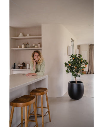 Een vrouw met krullend blond haar glimlacht, leunend op een aanrecht. Naast haar geven twee houten krukjes en een Kunst sinaasappelboom 120 cm een frisse sfeer aan de moderne, lichte keuken met planken en borden erachter.