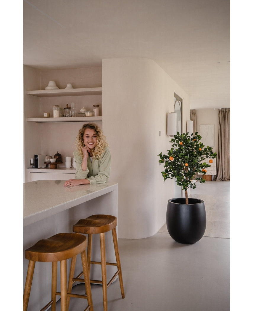 Een vrouw met krullend blond haar glimlacht, leunend op een aanrecht. Naast haar geven twee houten krukjes en een Kunst sinaasappelboom 120 cm een frisse sfeer aan de moderne, lichte keuken met planken en borden erachter.