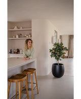 Een vrouw met krullend blond haar glimlacht achter een aanrecht met twee houten krukjes. De Kunst sinaasappelboom 120 cm in een pot voegt charme toe aan de moderne, minimalistische ruimte die is ingericht in neutrale tinten.