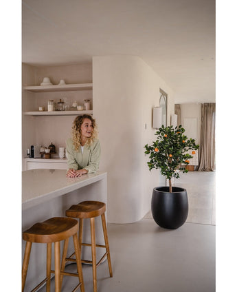 Een vrouw met krullend blond haar glimlacht achter een aanrecht met twee houten krukjes. De Kunst sinaasappelboom 120 cm in een pot voegt charme toe aan de moderne, minimalistische ruimte die is ingericht in neutrale tinten.