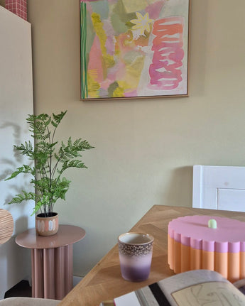 I et rum med pasteltema er der en 85 cm kunstig bregneplante på et lyserødt sidebord, en lilla og guld kop, en lyserød skål med en scalloped kant, en åben notesbog og på væggen et abstrakt maleri med grønne, pink og gule toner.