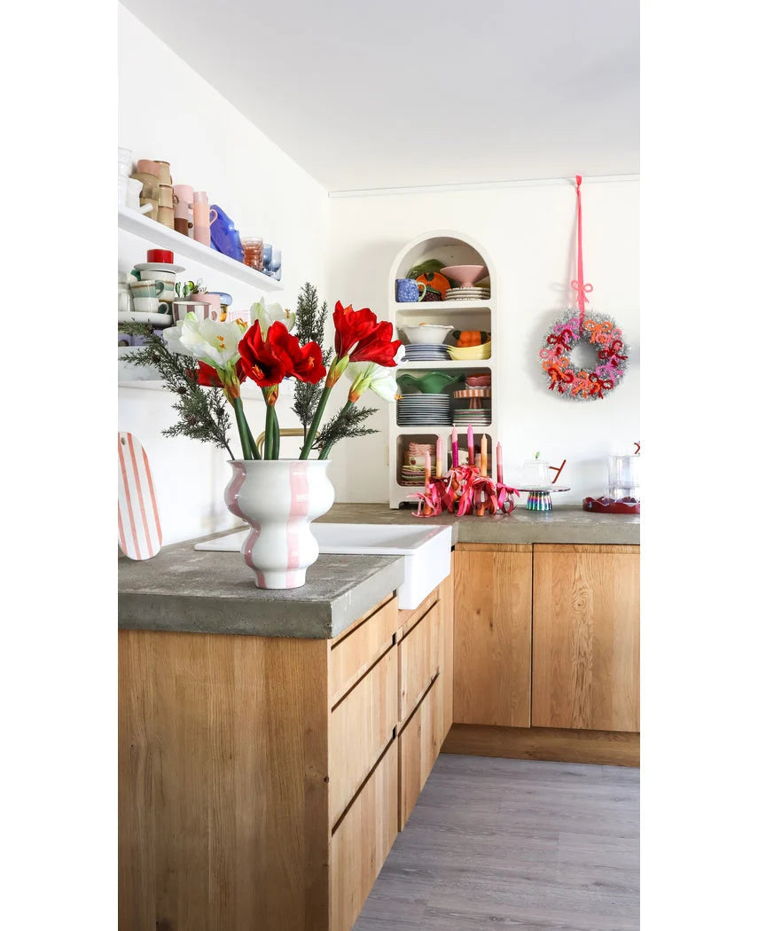 Een lichte keuken met houten kastjes, open witte planken met borden en potten, een feestelijke krans aan de muur en een Kunstbloem Amaryllis Real Touch rood 36 cm op het betonnen aanrecht.