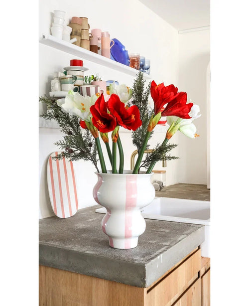Den Kunstige Blomst Amaryllis Real Touch rød 36 cm står i en hvid med pink stribet keramikvase på en betonbordplade i et lyst køkken med hylder med farverigt service i baggrunden.