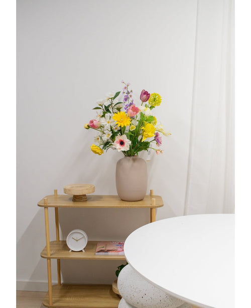 En lys træhylde viser en beige vase med Artificial Ranunculus Real Touch Yellow 60 cm blomster, et hvidt ur og en træbakke. En del af et rundt hvidt bord vises i forgrunden mod en almindelig hvid væg.