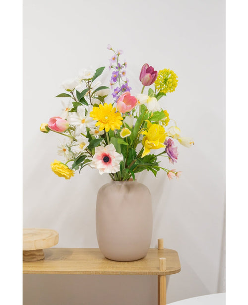 En beige vase med en Artificial Ranunculus Real Touch Yellow 60cm kunstig blomst står på et lyst træbord mod en almindelig hvid væg.
