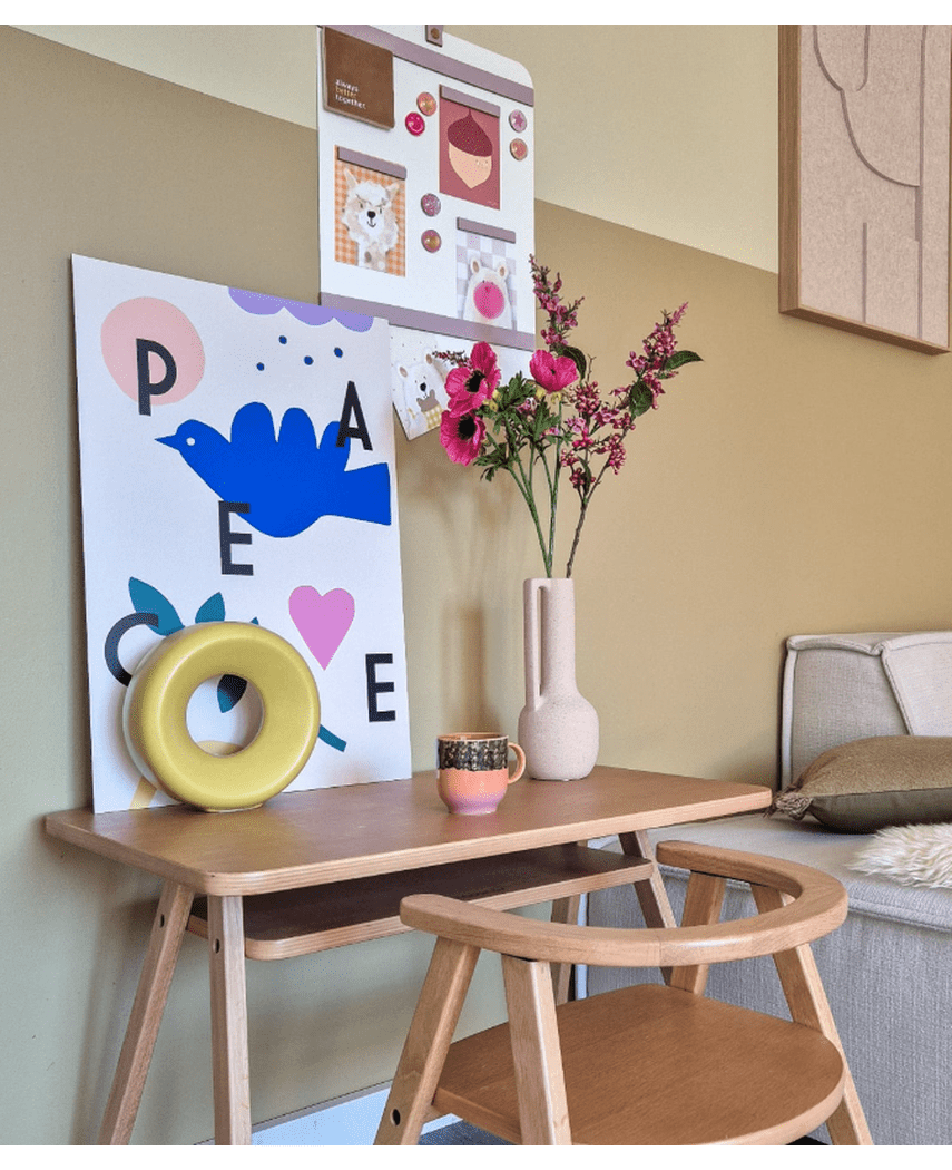 Een houten bureau met een bijpassende stoel toont een blauwe vogelafdruk, Kunstbloem Anemoon vertakt 56 cm roze, een rond geel ornament en een klein kopje. Boven hangt een prikbord met foto's en notities naast een beige bank.