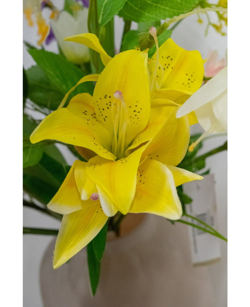 Et nærbillede af den kunstige blomst asiatisk lilje 66 cm gul, med knaldgule kronblade med mørke pletter, omgivet af grønne blade og andre blomster i en lys vase.