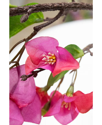 Close-up van Kunstbloem Bougainvillea 90 cm paars, met levendige paarse kunstbloemen met gele meeldraden en groene bladeren op een tak met bruine textuur, tegen een onscherpe witte achtergrond.