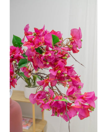 Een close-up van de Kunstbloem Bougainvillea 90 cm paars toont zijn levendige paarse bloemen en groene bladeren binnenshuis, met een potplant op een houten plank en een witte muur zacht vervaagd op de achtergrond.