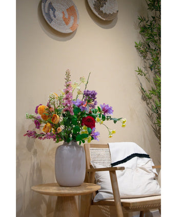 En hvid vase med Artificial Dogwood Real Touch Light Yellow 74cm står på et træsidebord ved siden af ​​en lys træstol med en hvid hynde og et tæppe med sorte kanter, fremhævet af vævet vægdekoration og en grøn plante i baggrunden.