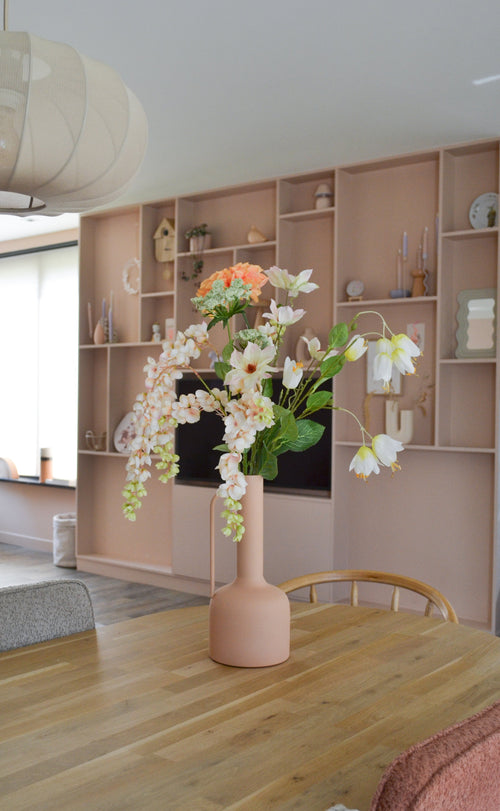 En moderne spisestue med et træbord med en ferskenfarvet vase indeholdende den kunstige blomst Fritillary 94 cm hvid. I baggrunden ses en stor indbygget reol dekoreret med stilfulde ting.