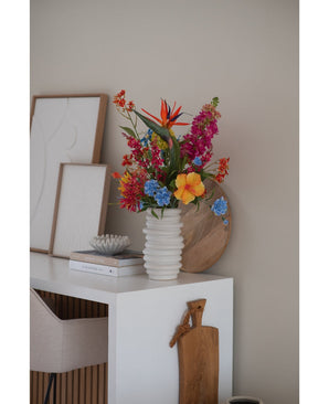 Een witte geribbelde vaas met Kunstbloem Kattenkruid 75 cm roze staat op een wit bureau met boeken, ingelijste kunst, een houten dienblad en een snijplank, naast een beige stoel in een moderne minimalistische kamer.
