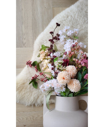 Den kunstige blomst Jilly 70 cm pink kunstig blomst står i en beige vase på et trægulv ved siden af ​​et blødt elfenbensfarvet fåreskindstæppe.