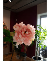 Et nærbillede af den kunstige blomsterpæon 61 cm blød pink, vist indendørs med grønne blade og potteplanter i baggrunden, badet i naturligt vindueslys.