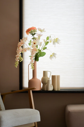 Et pastel stuehjørne med en træstol, en blød fodskammel og en vindueskarm med Artificial Flower Fritillary 94 cm hvid kunstig blomst i en lyserød vase ved siden af ​​minimalistisk keramisk indretning mod hvide persienner.