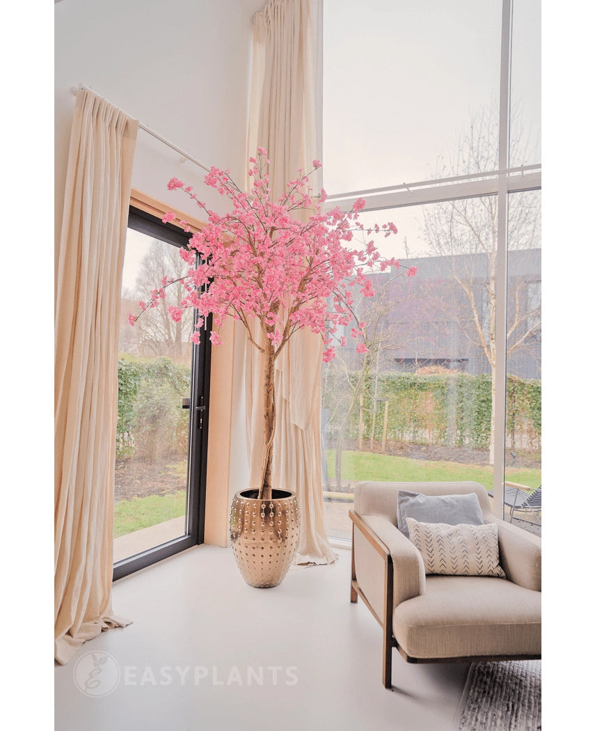 De Kunstboom Kersenbloesem roze 240 cm staat in een decoratieve pot bij een groot raam in een lichte, moderne woonkamer met crèmekleurige gordijnen, een beige fauteuil en uitzicht op de tuin buiten.