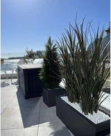 Kunstgræs Bordeaux UV 120cm i rektangulære plantekasser med hvide sten dekorerer en moderne udendørs terrasse mod en baggrund af blå himmel og træer i det fjerne.