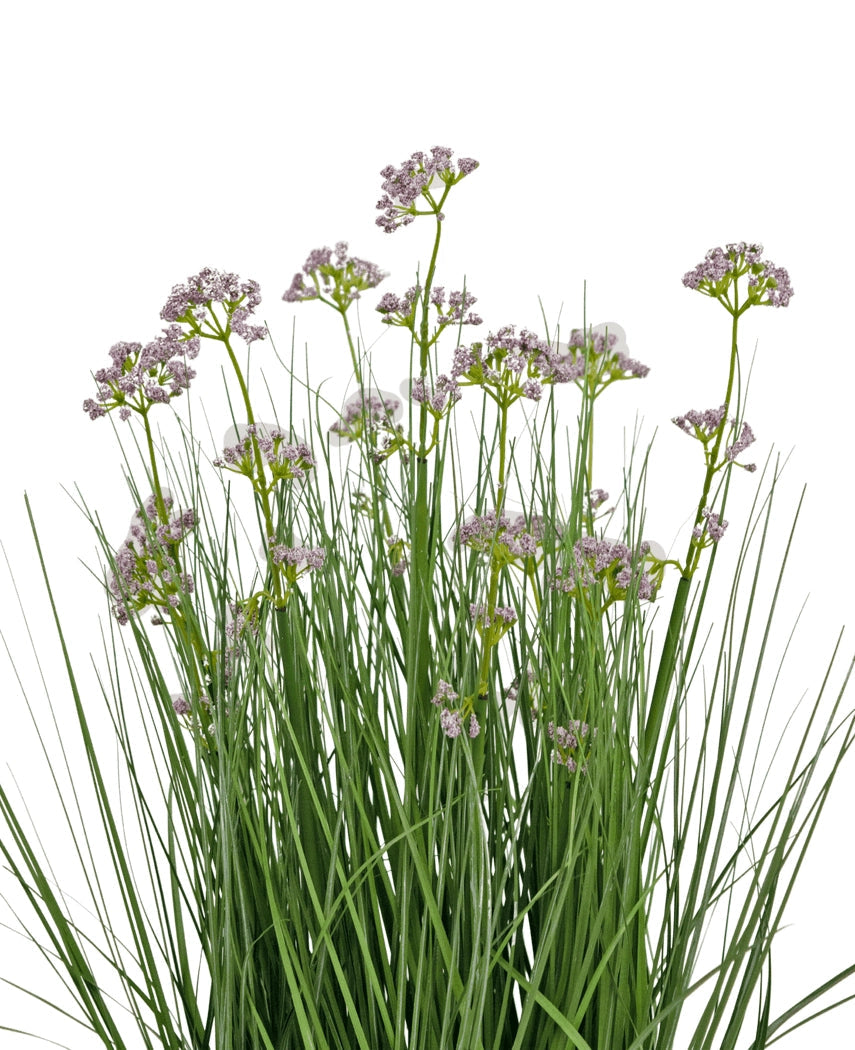 De Kunstgras Prei Bloem Ui Graspot 60cm heeft hoge, dunne groene bladeren en trossen kleine, delicate lichtpaarse bloemen op lange stelen. Het arrangement is brandvertragend en afgebeeld tegen een witte achtergrond.