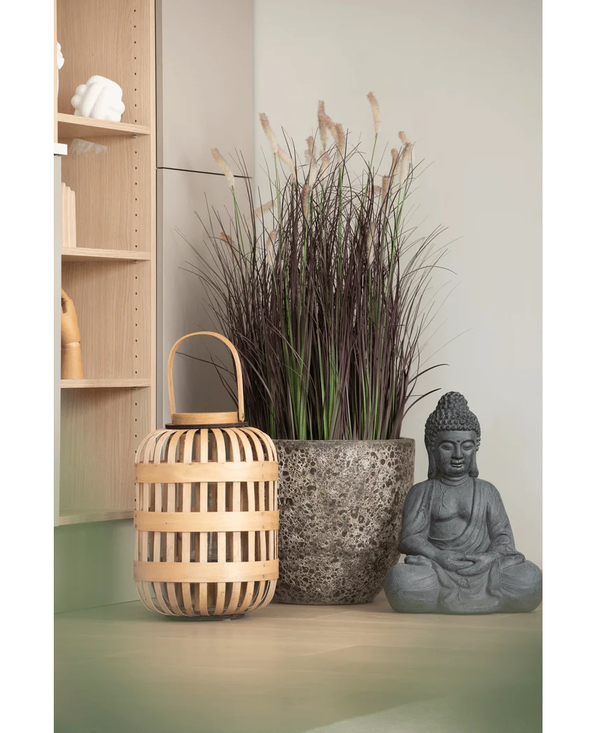 Een houten lantaarn, een hoge stenen pot met Kunstgras-uiengras-bonsai van 100 cm (brandvertragend) en een grijs Boeddhabeeld staan opgesteld op een lichtgekleurd oppervlak, vlakbij een houten plank en een bleke muur.