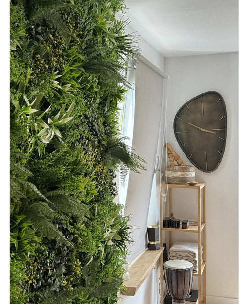 En frodig kunstig hæk Friskhed 100x100 cm UV lodret have dækker den ene væg i et lyst rum. I nærheden hænger et moderne trekantet vægur over en træhylde med håndklæder, en djembetromme, kurve og små dekorative stykker.