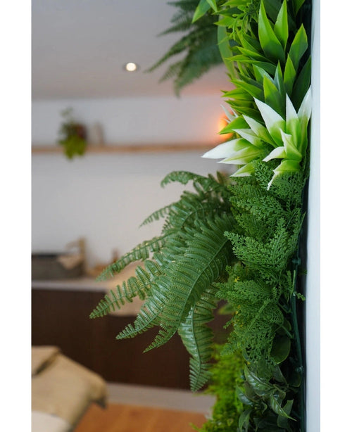 Et nærbillede af frodig grøn kunstig hæk Friskhed 100x100 cm UV kunstige planter pryder en lodret indervæg, med en sløret baggrund, der viser neutrale møbler og en lille hængende plante.