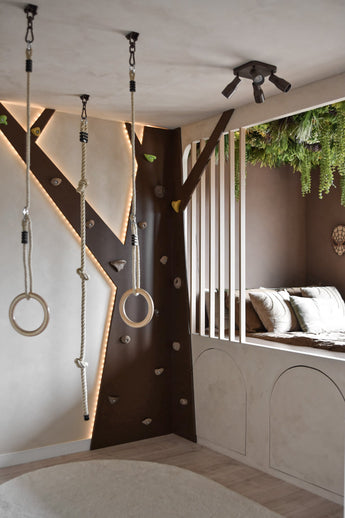 Een moderne kinderspeelkamer heeft een klimwand, gymringen, touw en een gezellige leeshoek met kussens en verticale latten. Kunsthaag Lush Deco Deluxe 100x100 cm UV voegt groen toe aan het plafond en vult de neutrale tinten en speelse verlichting van de kamer aan.