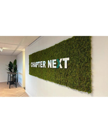 Een luxe interieuraccent: een groene wanddisplay met Kunsthaag Nordic Mos 50x50 cm UV-kaders, met "CHAPTER NEXT" in vette 3D-letters. Een tafel versierd met planten maakt het geraffineerde tafereel compleet.