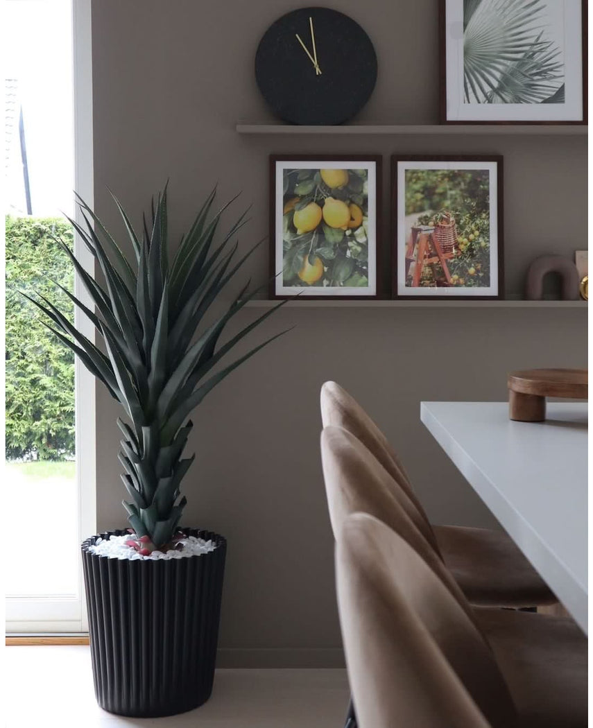 Een moderne eethoek met beige stoelen, een witte tafel, een wandklok en een plank met ingelijste foto's van citroen en bladeren. De grote Kunstplant Agave Pandora UV 120 cm voegt groen toe. Natuurlijk licht valt binnen vanaf de linkerkant.
