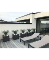 Een moderne patio heeft twee grijze loungestoelen en vijf vierkante plantenbakken, elk met een Kunstplant Agava Pandora UV 85 cm, tegen een wit met zwart gepaneelde muur voor een strakke, minimalistische look.