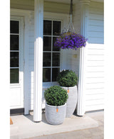 Een witte veranda van een huis heeft twee Kunstplant Buxus op stam romeinse stijl UV 65cm topiary plantenbakken met groene struiken, een hangende mand met paarse bloemen erboven, witte zuilen en een deur met glazen panelen aan de linkerkant.